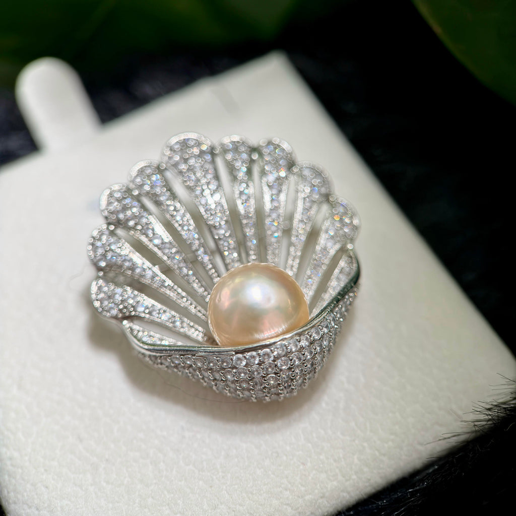 A shell-shaped silver ring brooch or pendant featuring a central pearl and pavé-set clear stones, displayed on a white jewelry stand.