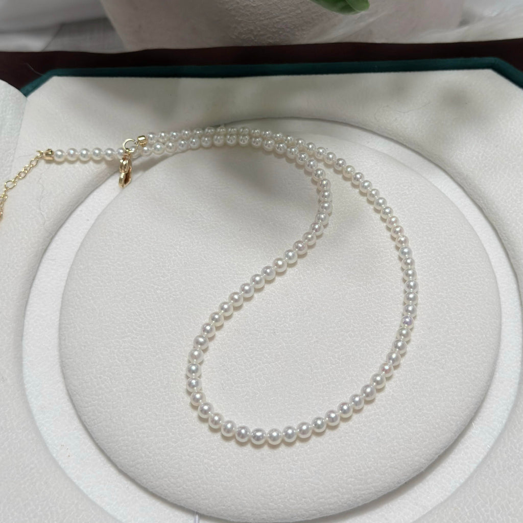 A delicate strand of white freshwater pearls with a gold clasp, displayed on a white circular jewelry stand.