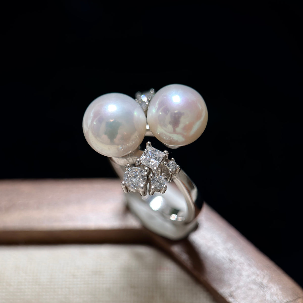 A silver ring featuring two large round white pearls and a cluster of small clear stones, displayed on a beige surface against a black background.
