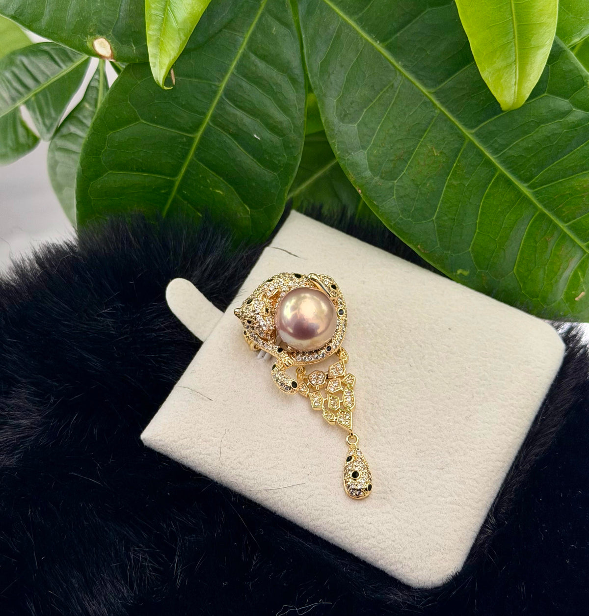 A gold-toned brooch featuring a central pink pearl and delicate crystal detailing, displayed on a white jewelry cushion with green leaves and a black faux-fur surface in the background.