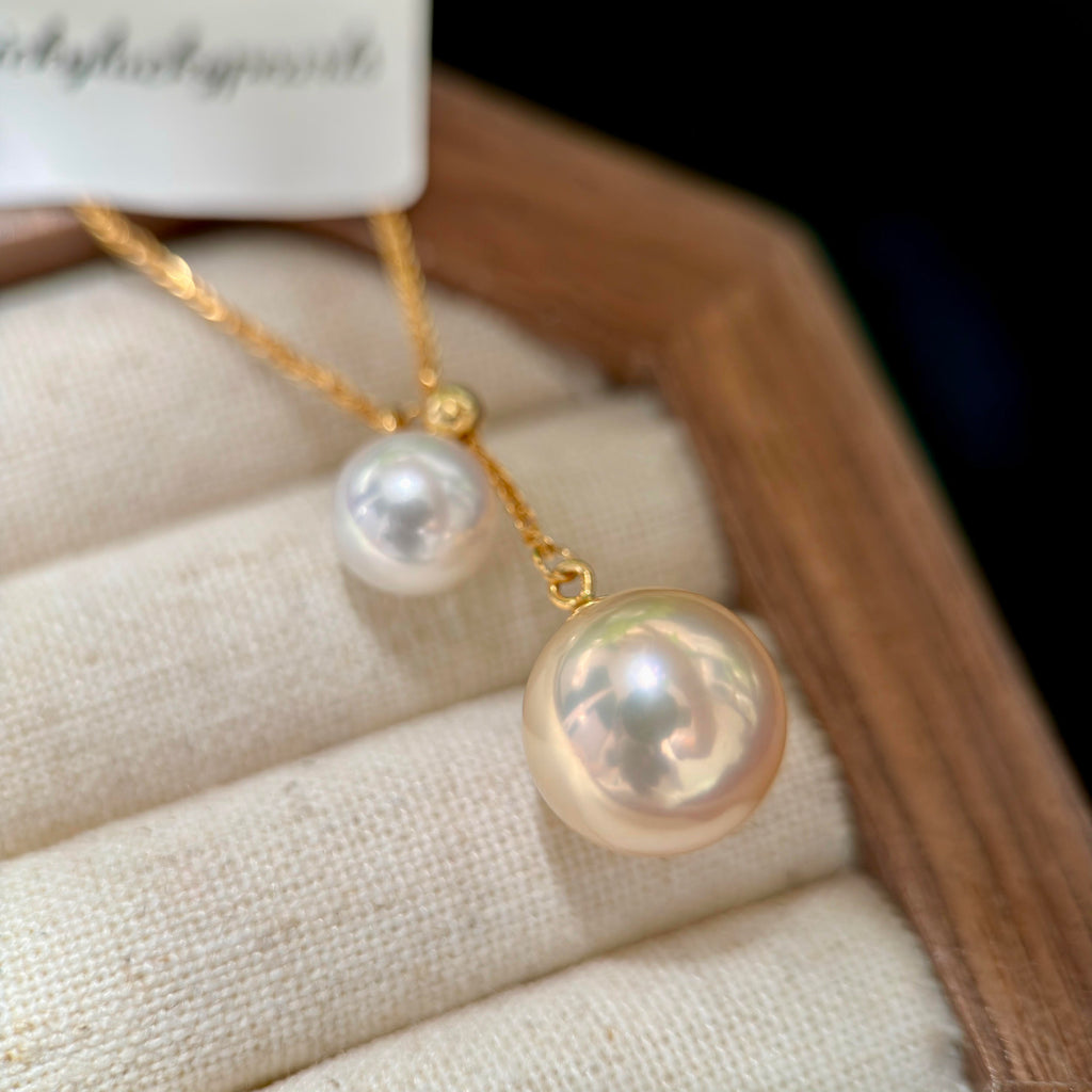 A gold necklace with two pearl-like drops, one larger pearlescent drop and one smaller drop, displayed on a cream fabric jewelry cushion inside a wooden box.