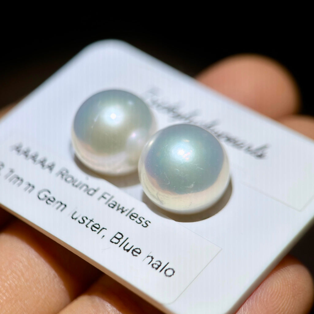 Two round iridescent freshwater pearls on a white display card held in a hand. The card reads 'AAAAA Round Flawless 11mm Gem oster, Blue halo'.