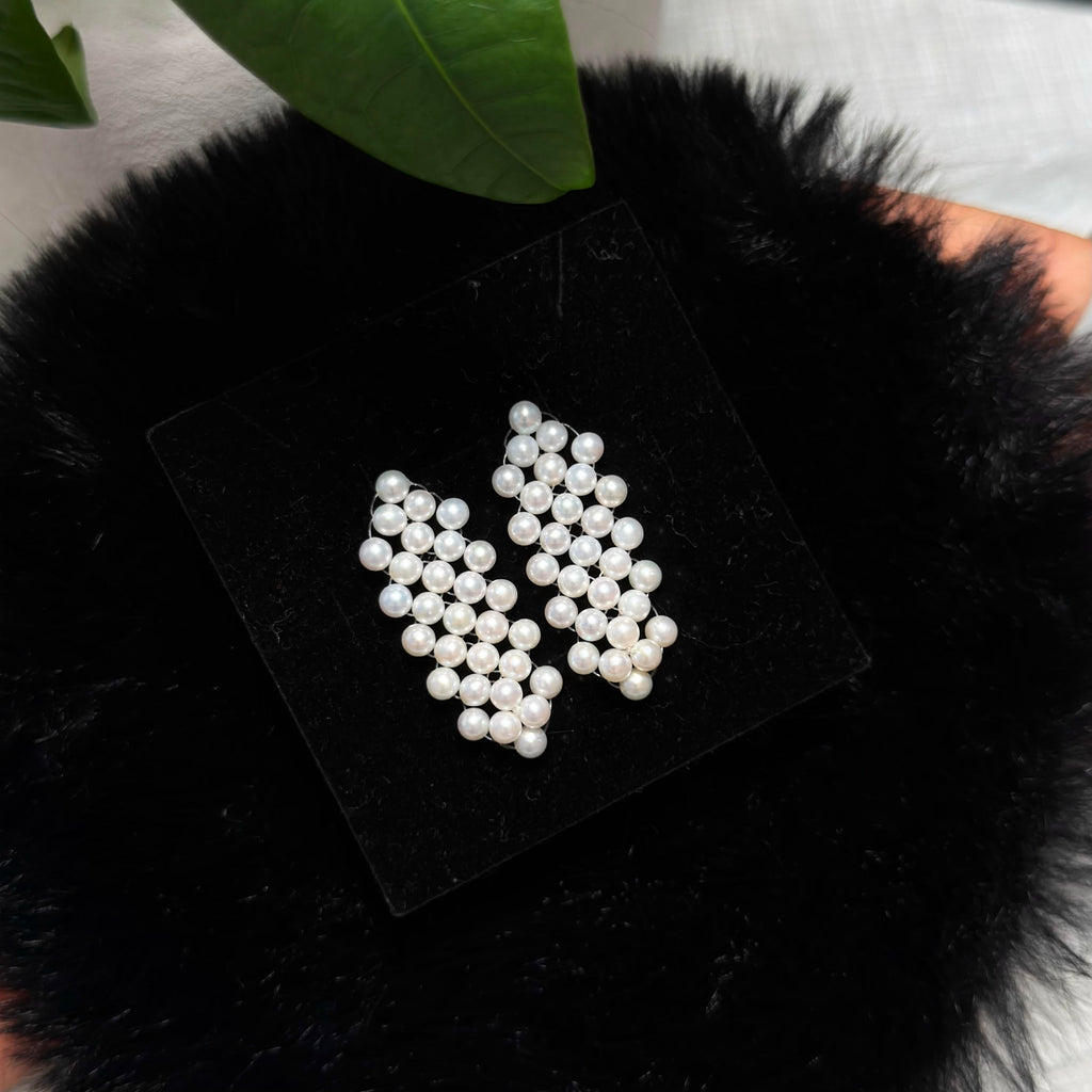 A pair of white pearl stud earrings arranged on a black square display card, placed on a black fluffy surface with a green leaf in the background.