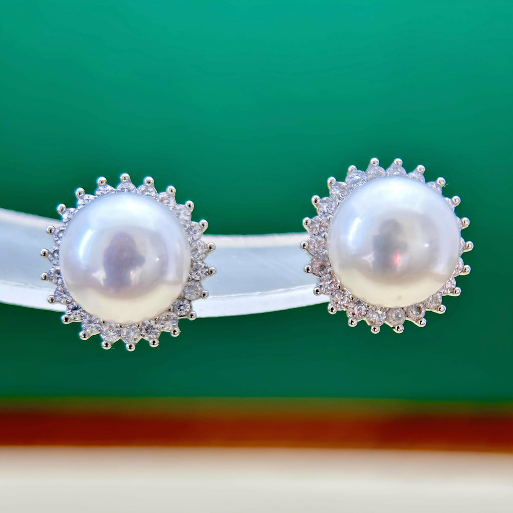 A pair of silver-toned stud earrings with large white pearls surrounded by small clear stones, displayed on a clear stand against a green background.