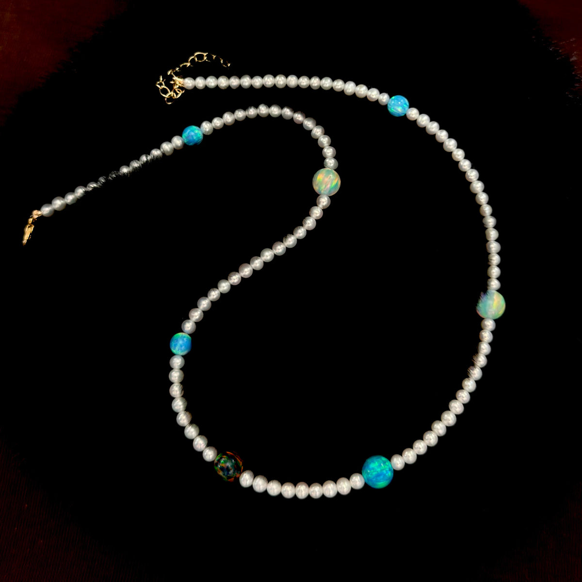 A beaded necklace made of small white pearls with several round blue and iridescent opal-like beads, plus a small multicolor decorative bead and gold-tone clasp, photographed on a black background.