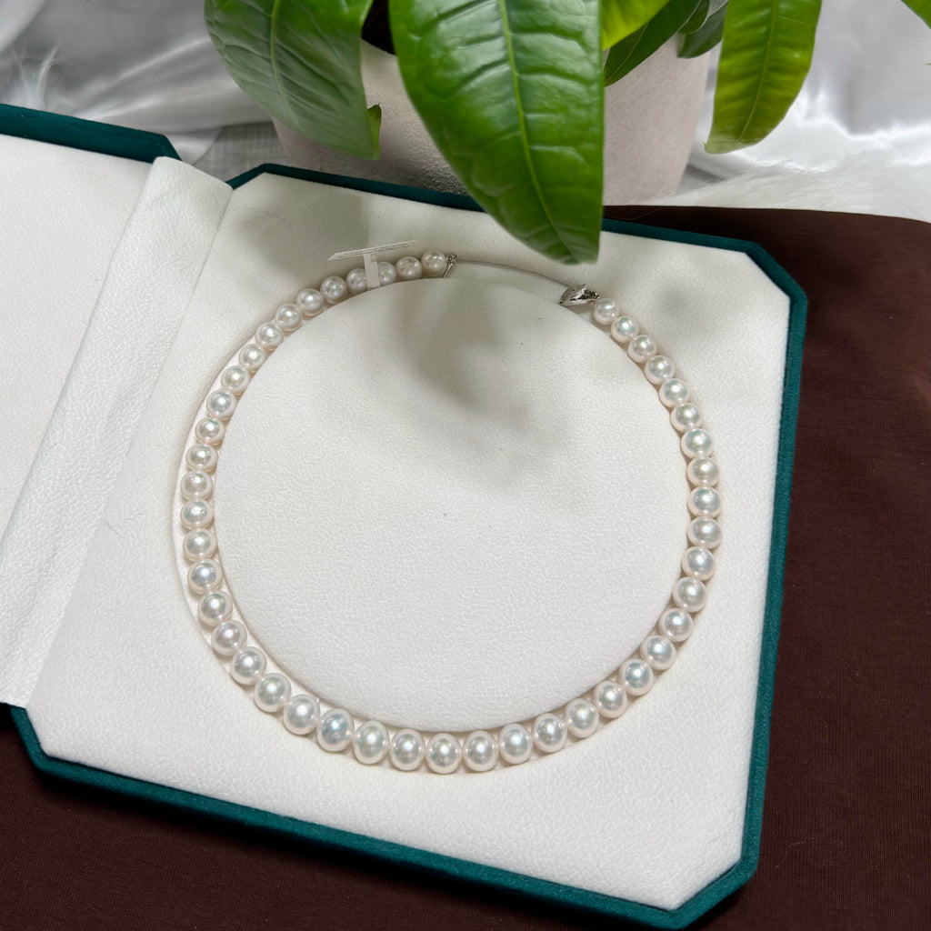A classic freshwater pearl necklace arranged in a circle inside an open teal jewelry box with a white insert, with green leaves hanging over the top.