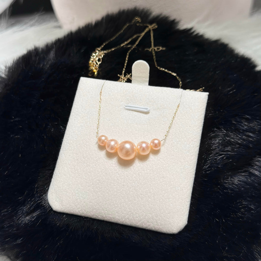 A gold chain necklace with five peach-pink pearls of varying sizes, displayed on a white jewelry stand against a black faux fur background.