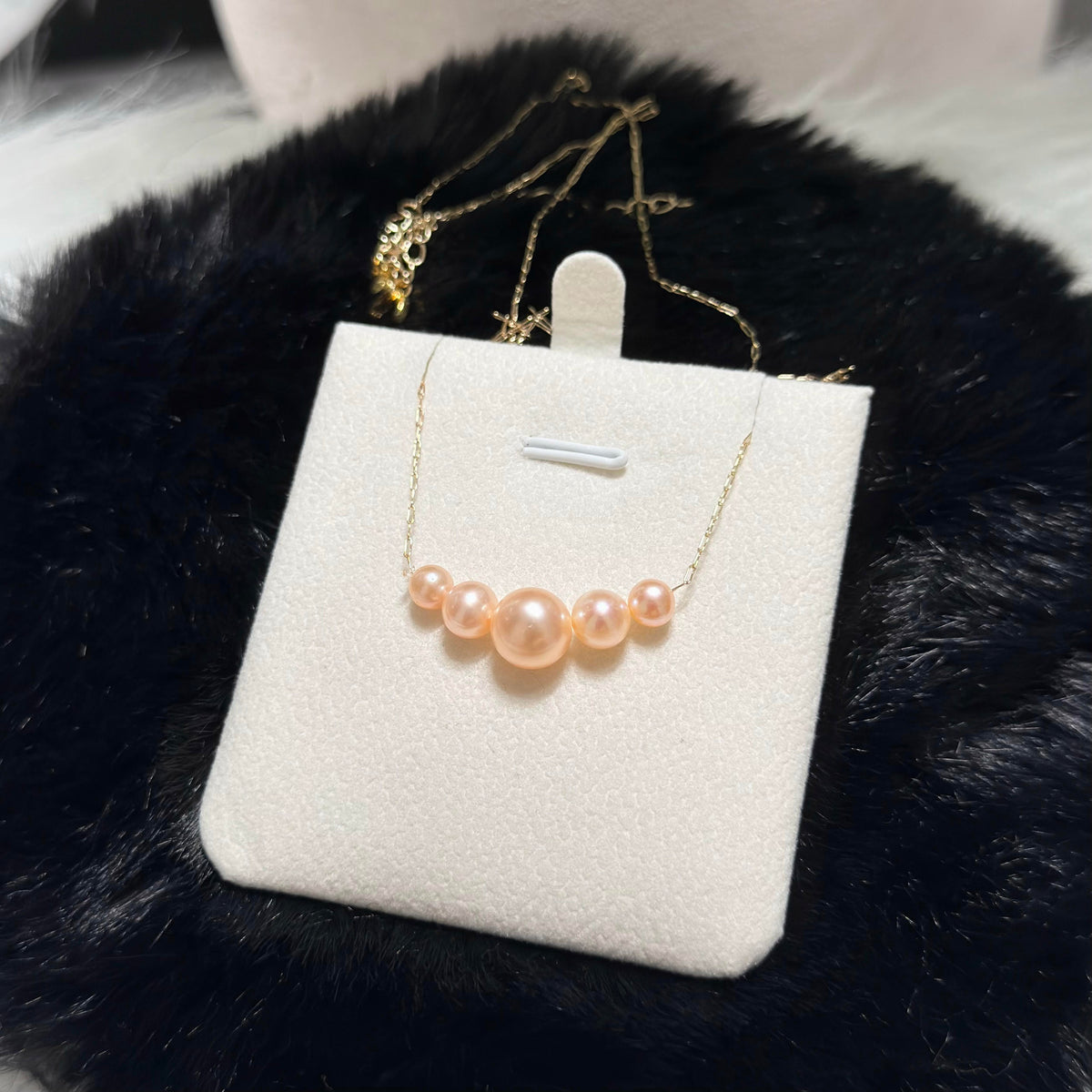 A gold chain necklace with five peach-pink pearls of varying sizes, displayed on a white jewelry stand against a black faux fur background.