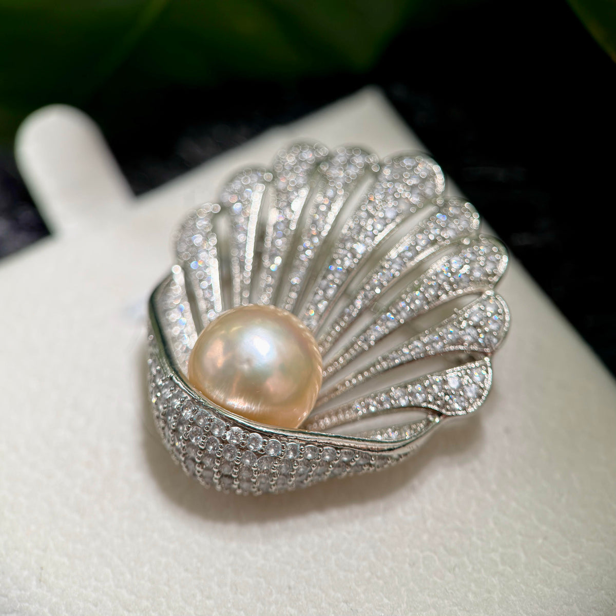 A silver shell-shaped ring featuring a large, lustrous pearl at the center and pavé-set clear stones along the outer flaps, displayed on a white jewelry stand.