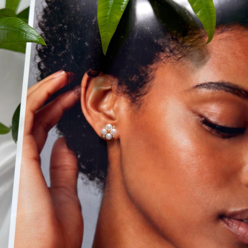 A close-up lifestyle shot of a model wearing a small cluster stud earring made of white pearls, with green leaves framing the top of the photo.
