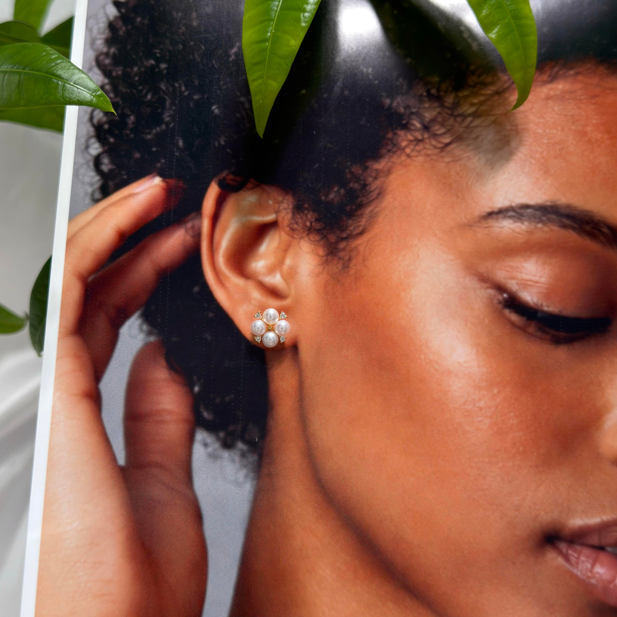 A close-up lifestyle shot of a model wearing a small cluster stud earring made of white pearls, with green leaves framing the top of the photo.
