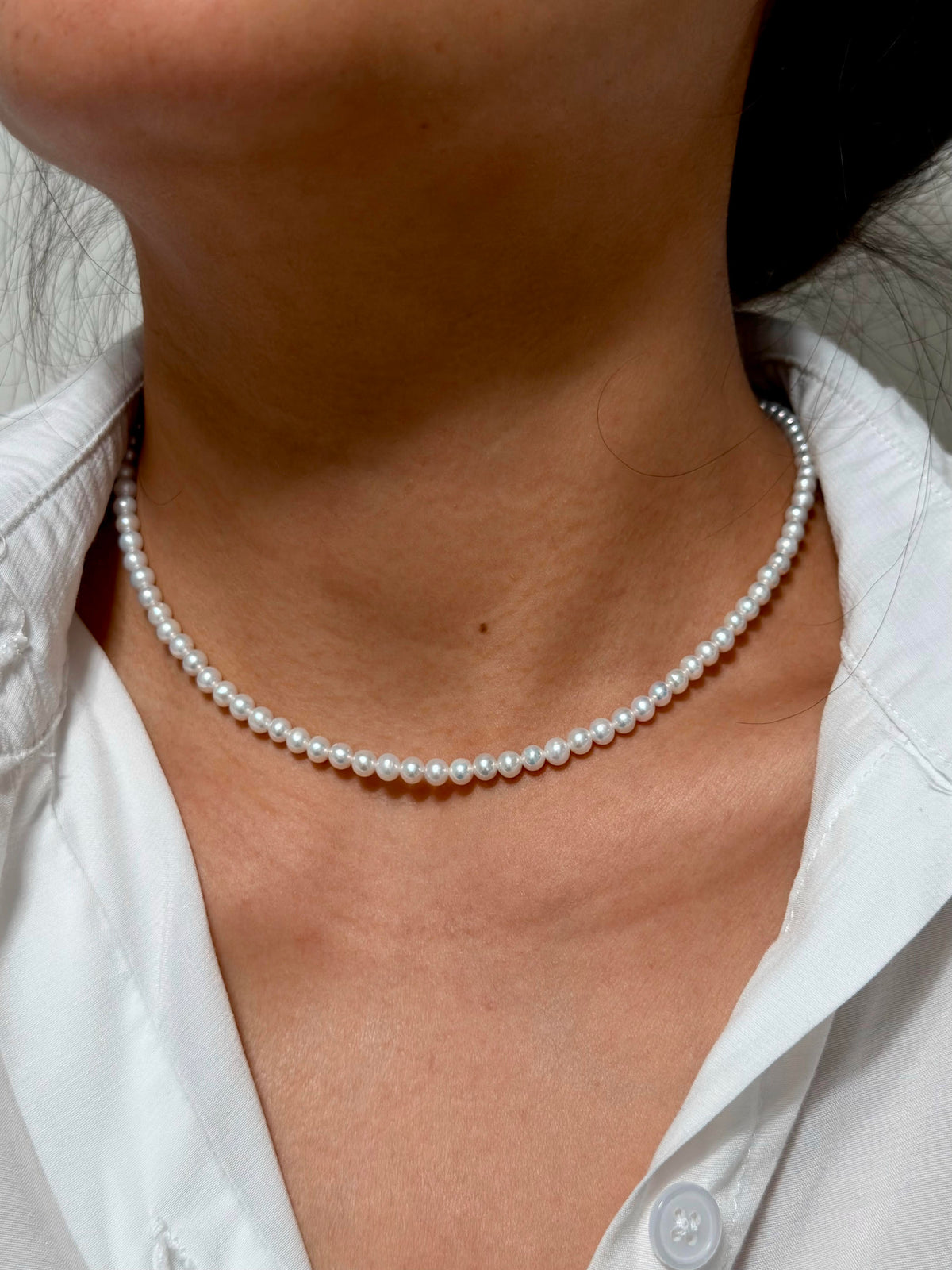 A close-up of a person wearing a delicate single-strand pearl choker necklace with a white button-down shirt.