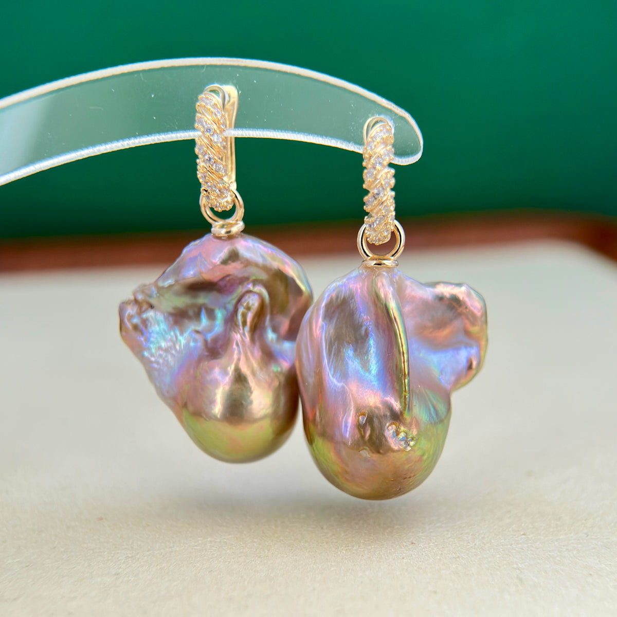 A pair of gold hoop earrings with large, baroque-shaped iridescent freshwater pearls, displayed on a clear plastic stand against a green and beige background.