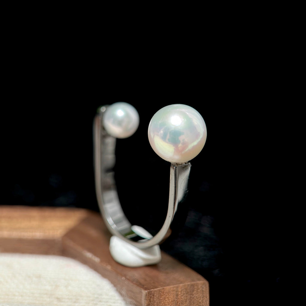A silver-toned open ring earring with two round white pearls, displayed on a small stand against a black background.