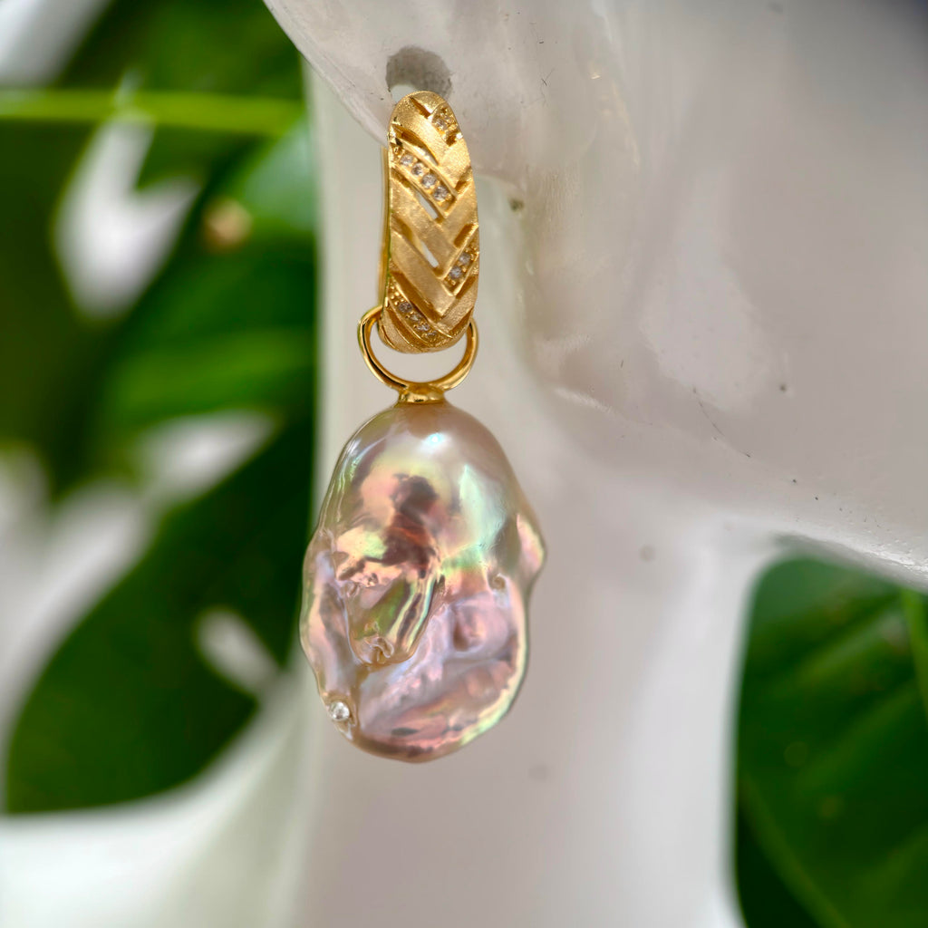 A gold drop earring featuring a large, irregular baroque pearl with iridescent pink and green hues, displayed on a white ceramic stand with green leaves in the background.