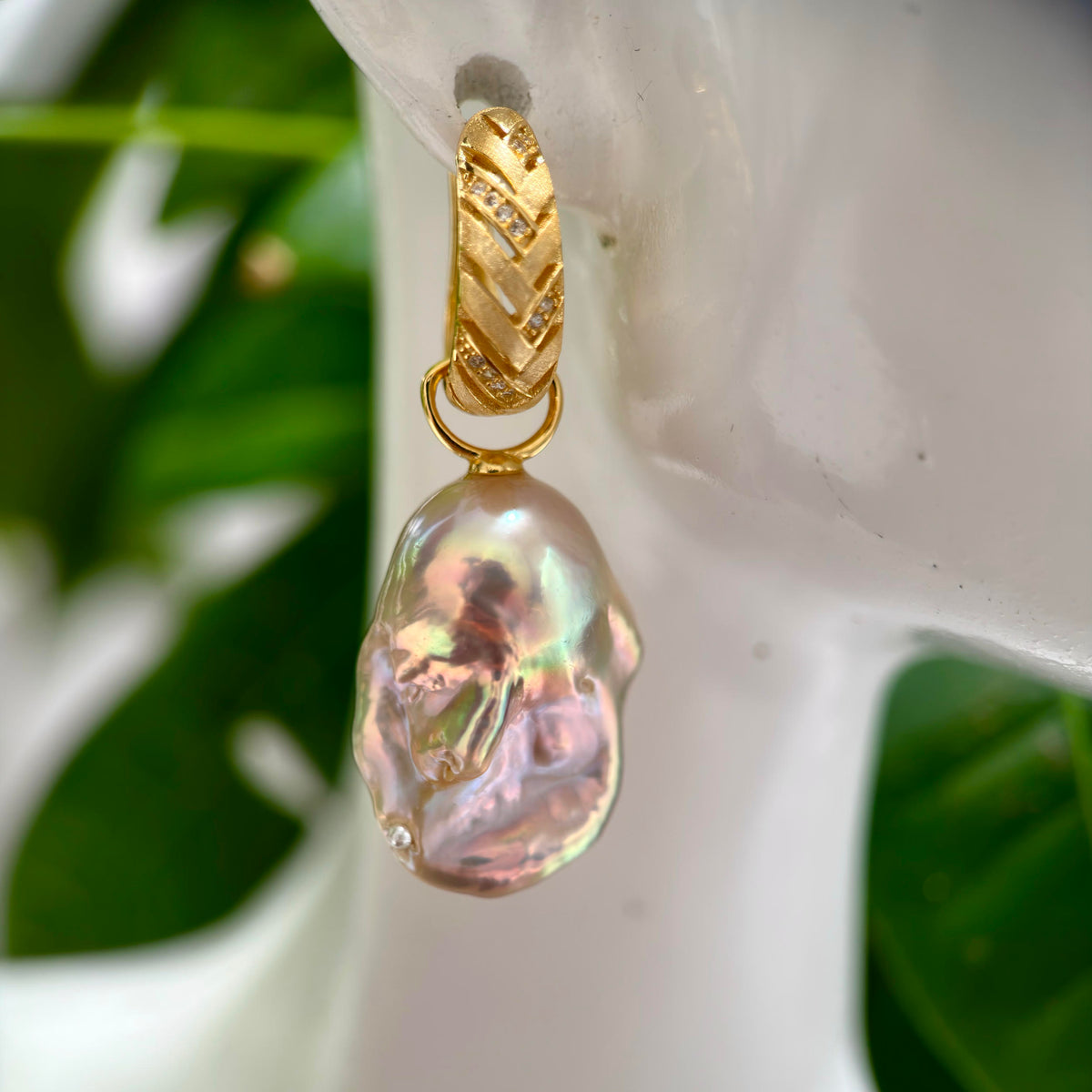 A gold drop earring featuring a large, irregular baroque pearl with iridescent pink and green hues, displayed on a white ceramic stand with green leaves in the background.