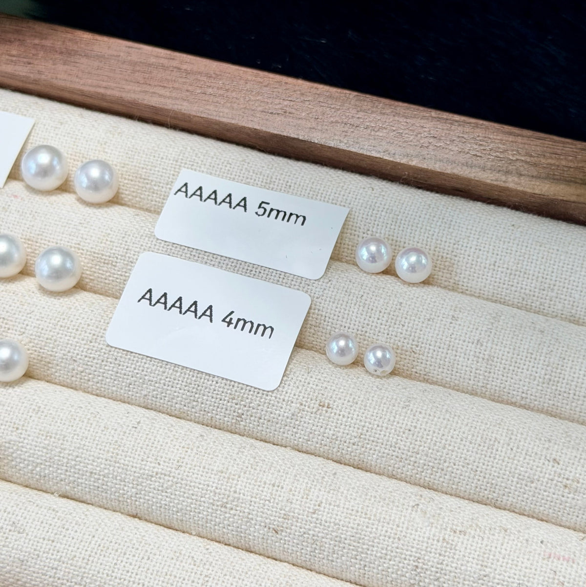 White freshwater pearls displayed on a beige fabric jewelry tray, with two white paper labels reading 'AAAAA 5mm' and 'AAAAA 4mm' placed over the pearls.