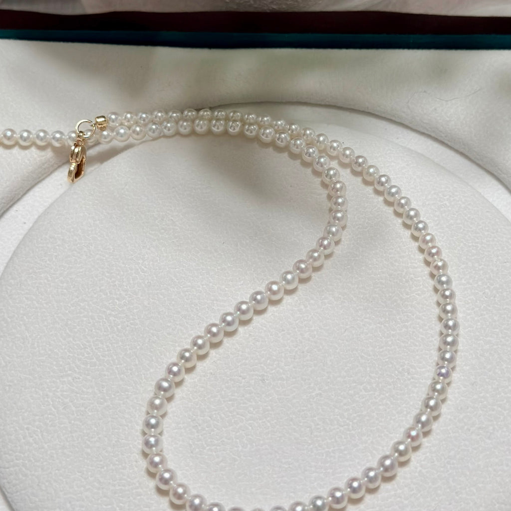 A delicate strand of white freshwater pearls draped in an S-shape on a white circular jewelry display, featuring a small gold clasp at one end.