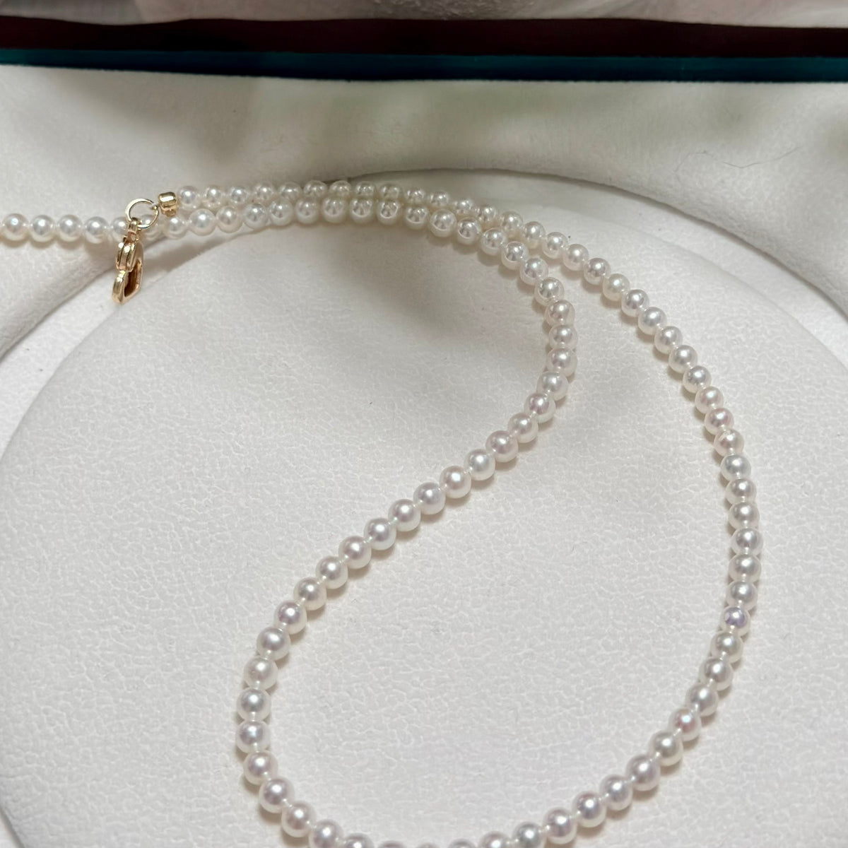 A delicate strand of white freshwater pearls draped in an S-shape on a white circular jewelry display, featuring a small gold clasp at one end.