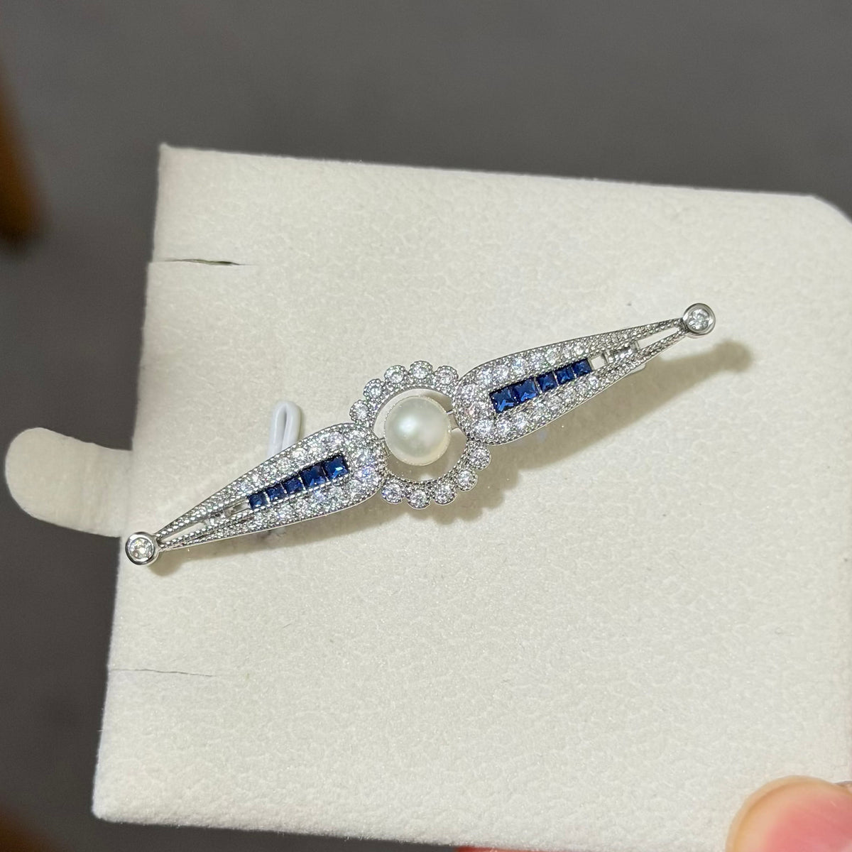 An Art Deco style brooch featuring a central white pearl surrounded by clear pavé stones, with blue stone accents on either side, displayed on a cream jewelry stand.