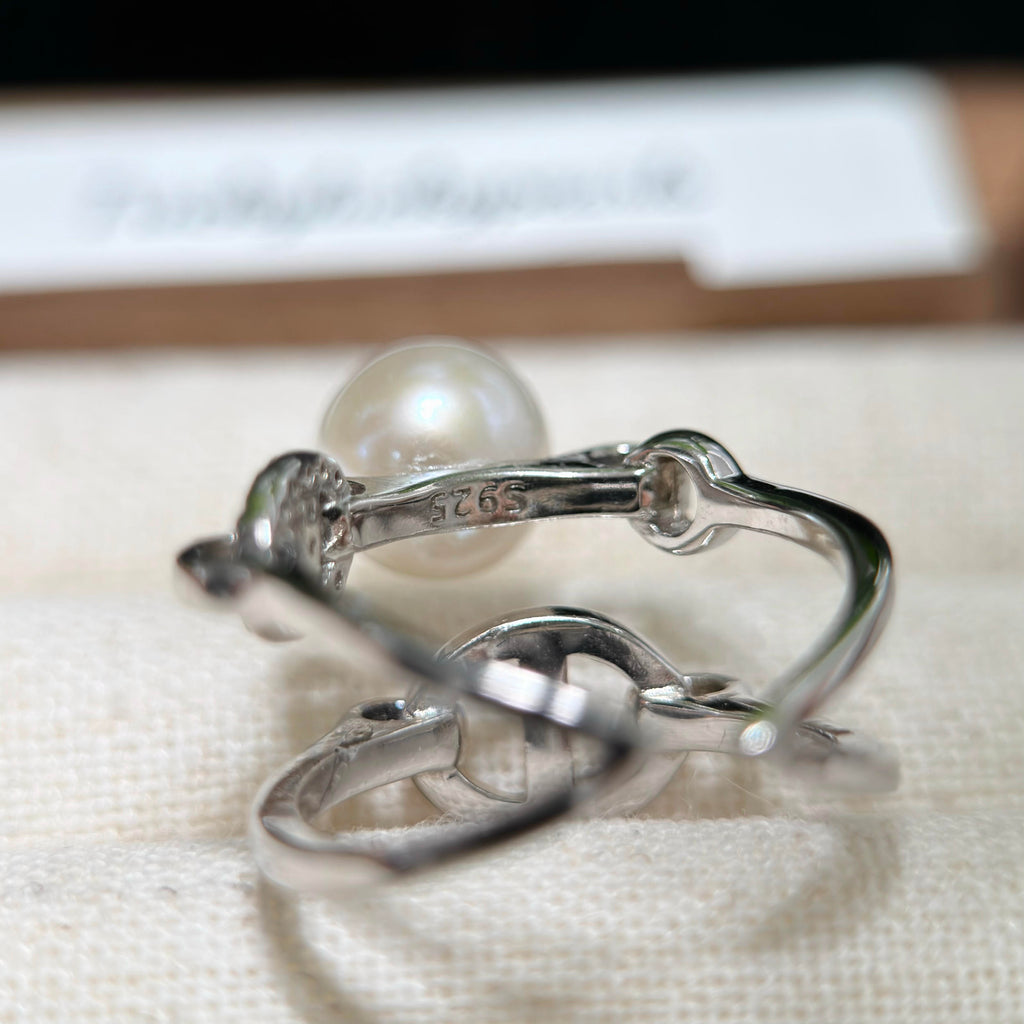 A sterling silver ring with a single round white pearl, shown close-up on a textured cream fabric surface with a blurred card in the background.