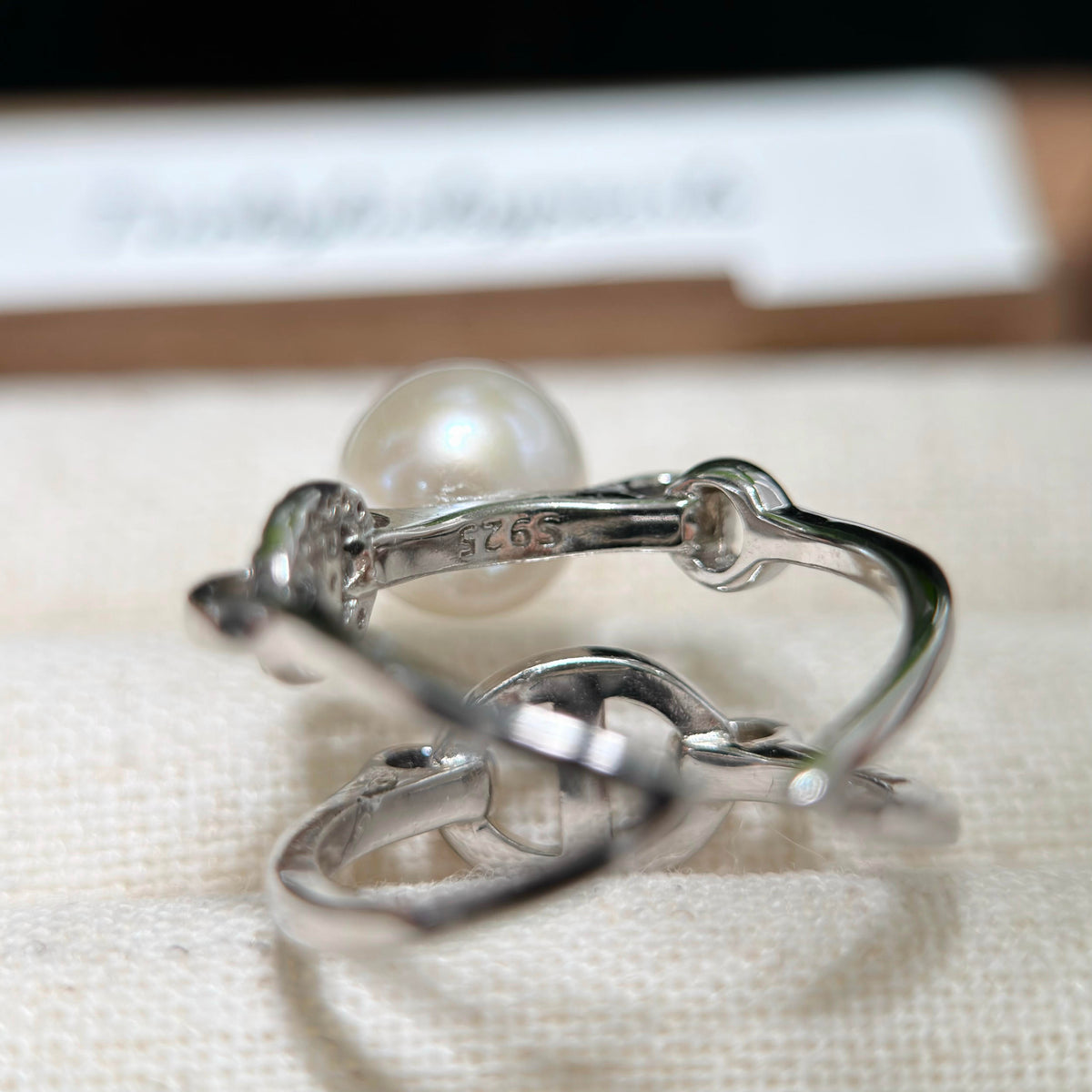 A sterling silver ring with a single round white pearl, shown close-up on a textured cream fabric surface with a blurred card in the background.