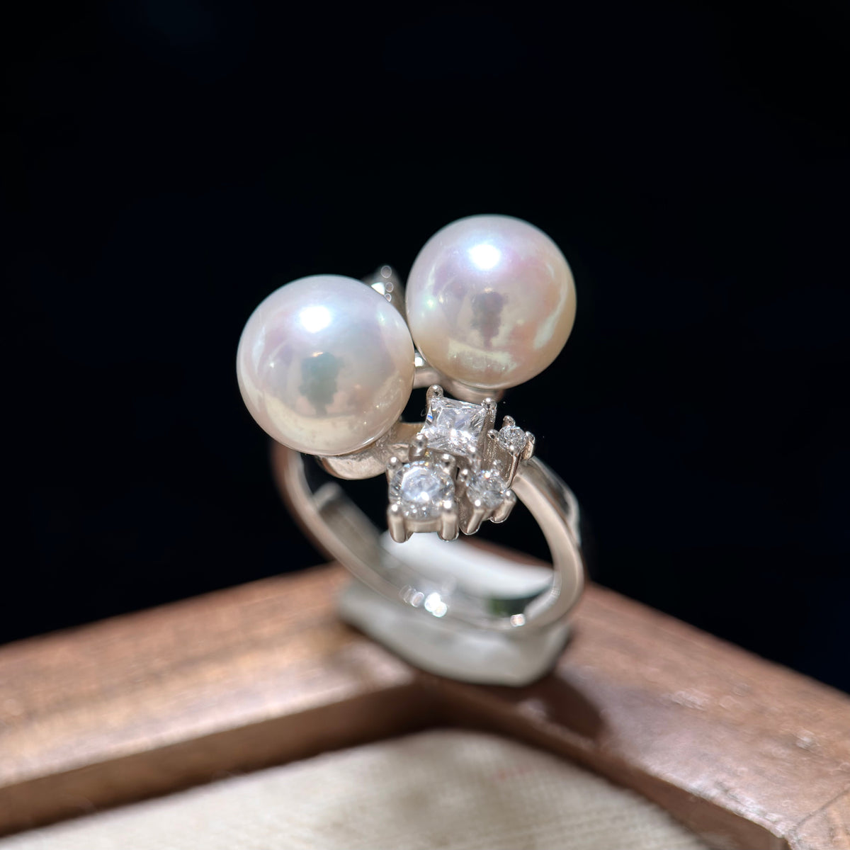 A silver ring featuring two large white pearls and a small cluster of clear stones, displayed on a wooden surface against a dark background.
