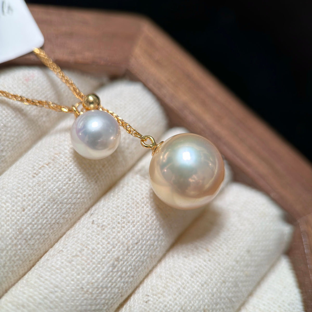 A close-up of a gold necklace with two white freshwater pearls, one larger and one smaller, displayed on a cream fabric jewelry stand.