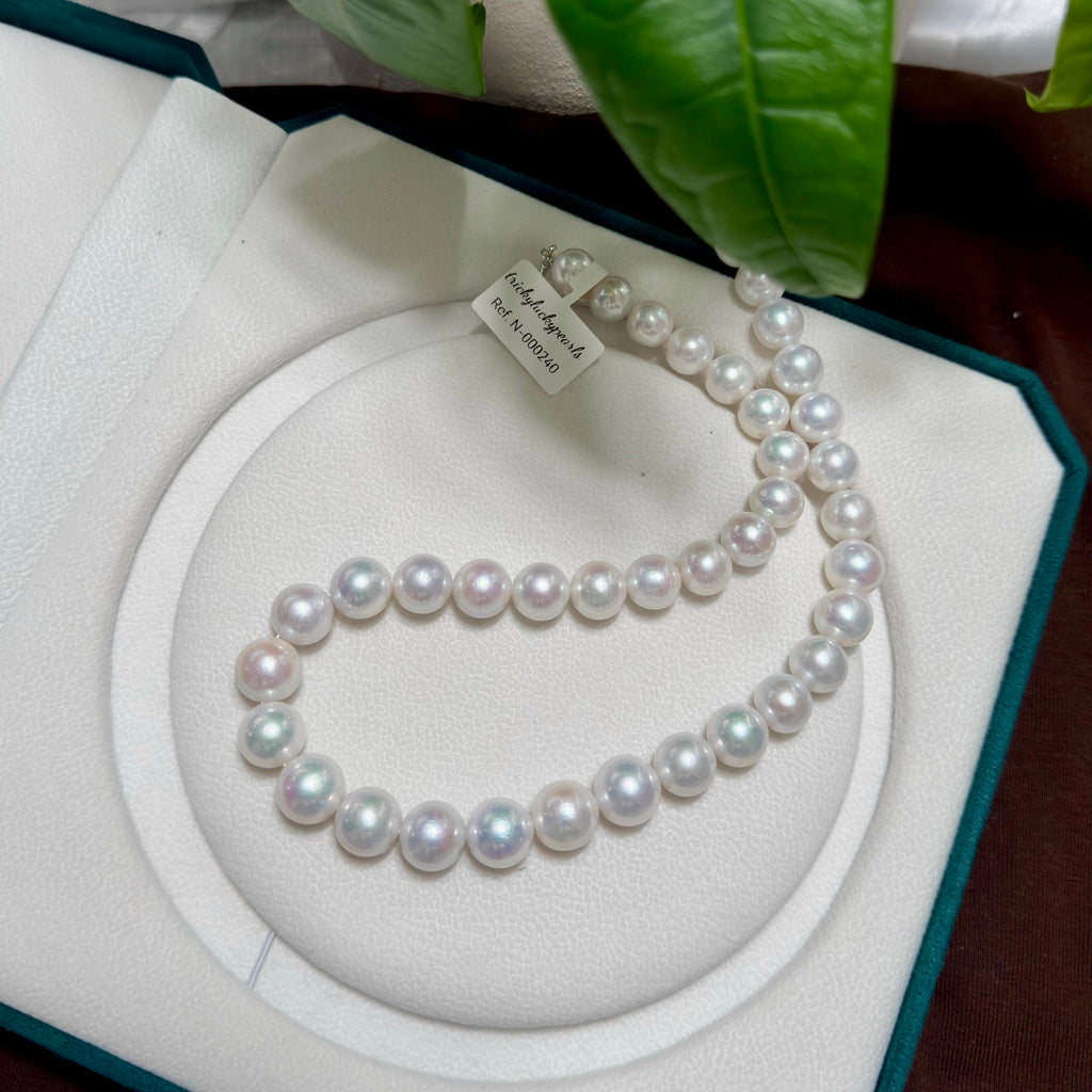 A strand of freshwater pearl necklace displayed inside an open jewelry box, with a small paper tag attached. The pearls are white with subtle iridescent pinks and greens, arranged on a cream-colored circular insert.