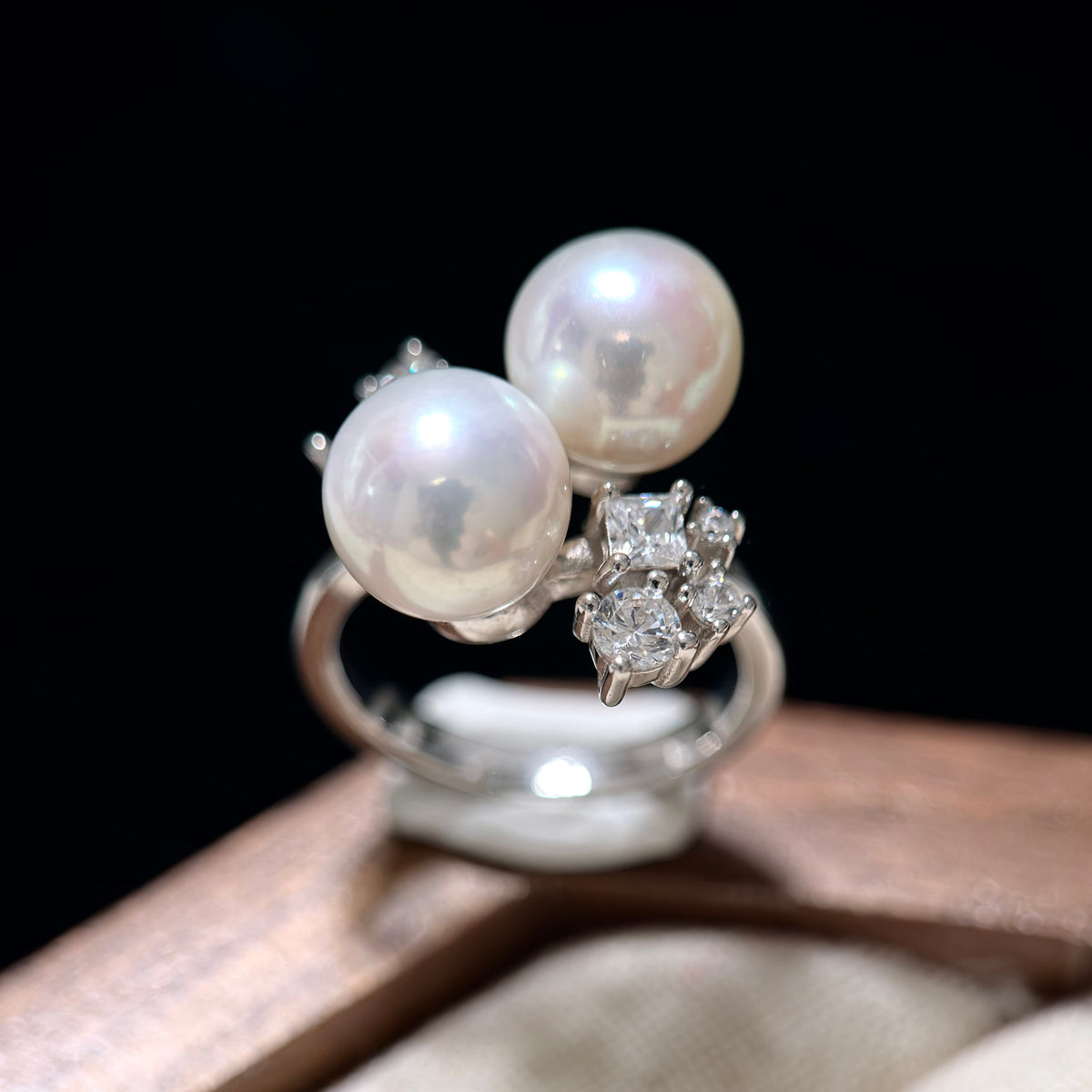 A silver ring featuring two large white pearls and a cluster of clear gemstones, displayed on a wooden surface against a black background.