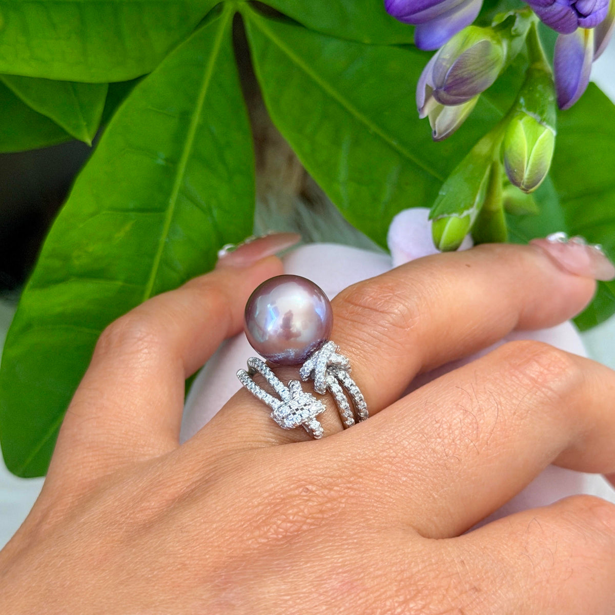 A close-up of a hand wearing a silver ring with a large round pearl and a knot design paved with small clear stones, set against green leaves and purple flowers.