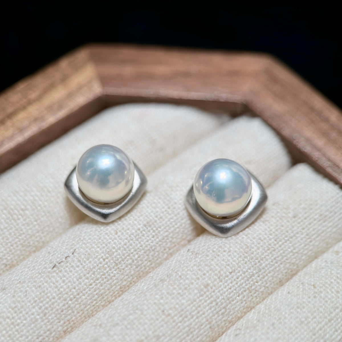 A pair of silver stud earrings with round white pearls set in a square bezel, displayed on a cream fabric ring cushion inside a wooden box.