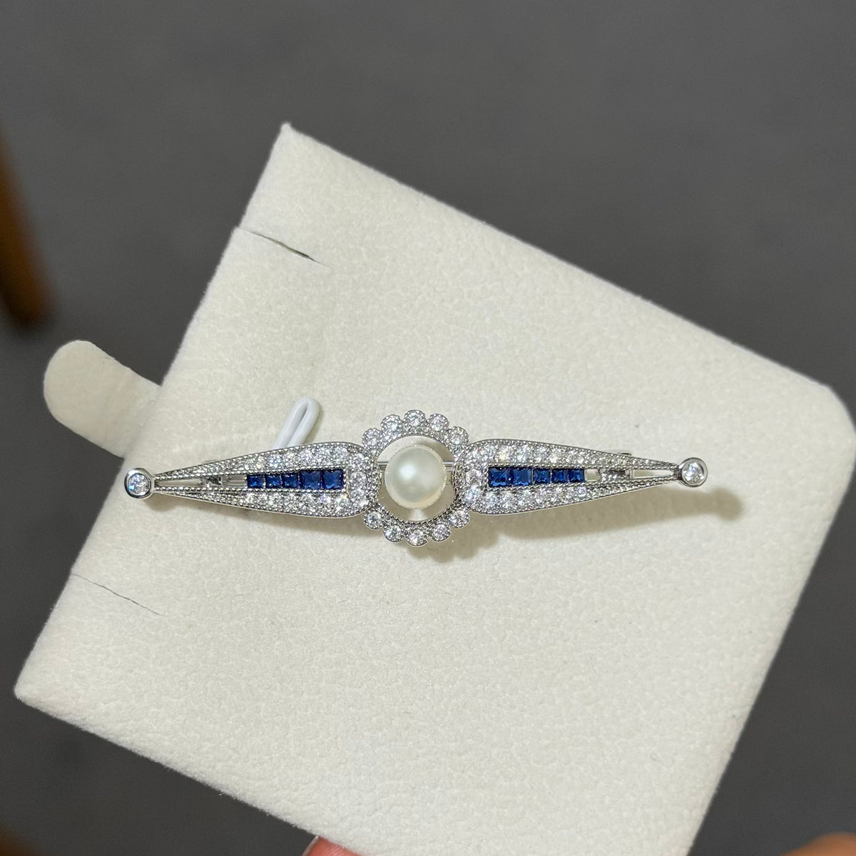 An Art Deco style silver-toned brooch featuring a central white pearl surrounded by small clear stones, with blue stone accents on either side, displayed on a cream jewelry box.