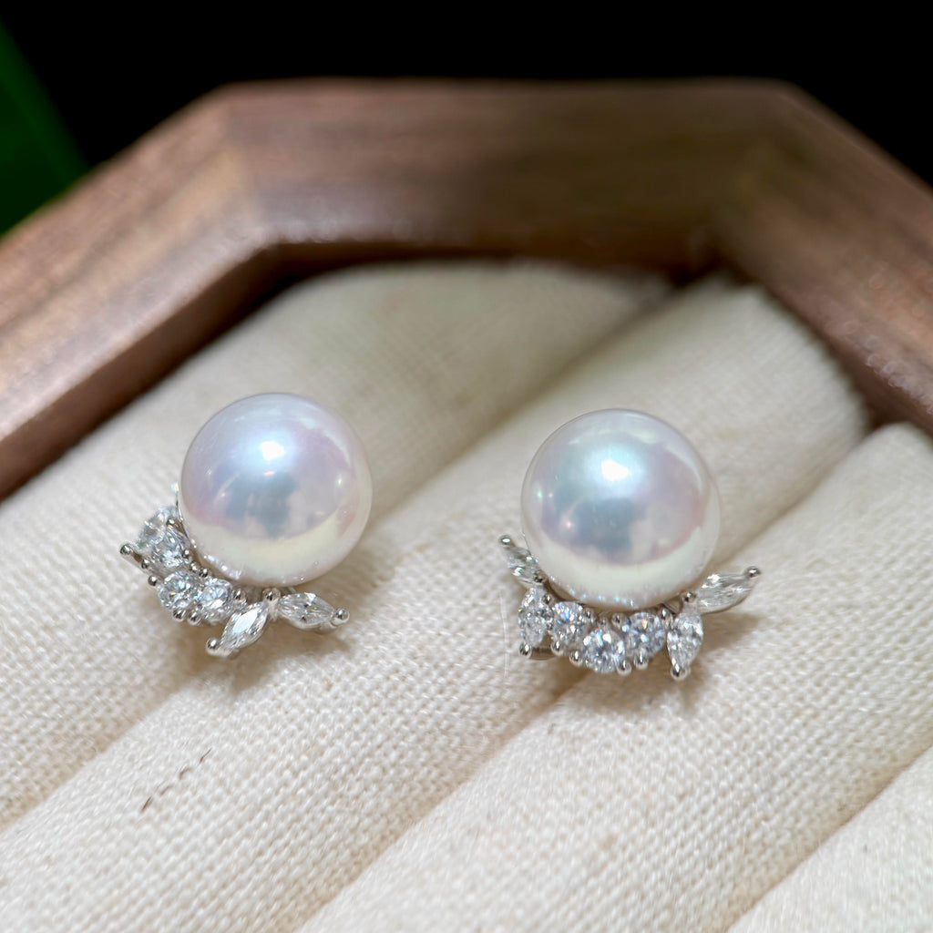 A pair of pearl stud earrings with silver-toned floral settings and clear marquise and round stones, displayed on a cream fabric surface inside a wooden box.