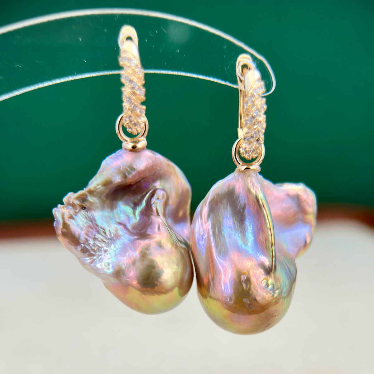 A pair of baroque pearl drop earrings with gold-toned twisted hooks and small clear stones, hanging against a dark green and light beige background.
