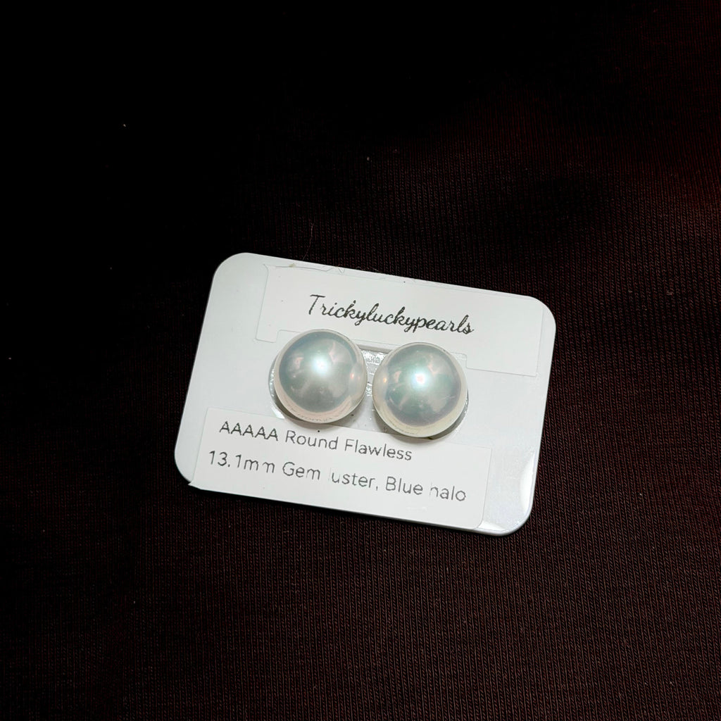 A pair of round flawless blue nalo pearls on a white display card by TrickyLuckyPearls, set against a dark brown textured background.