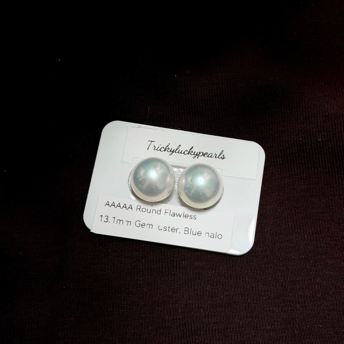 A pair of round flawless blue nalo pearls on a white display card by TrickyLuckyPearls, set against a dark brown textured background.