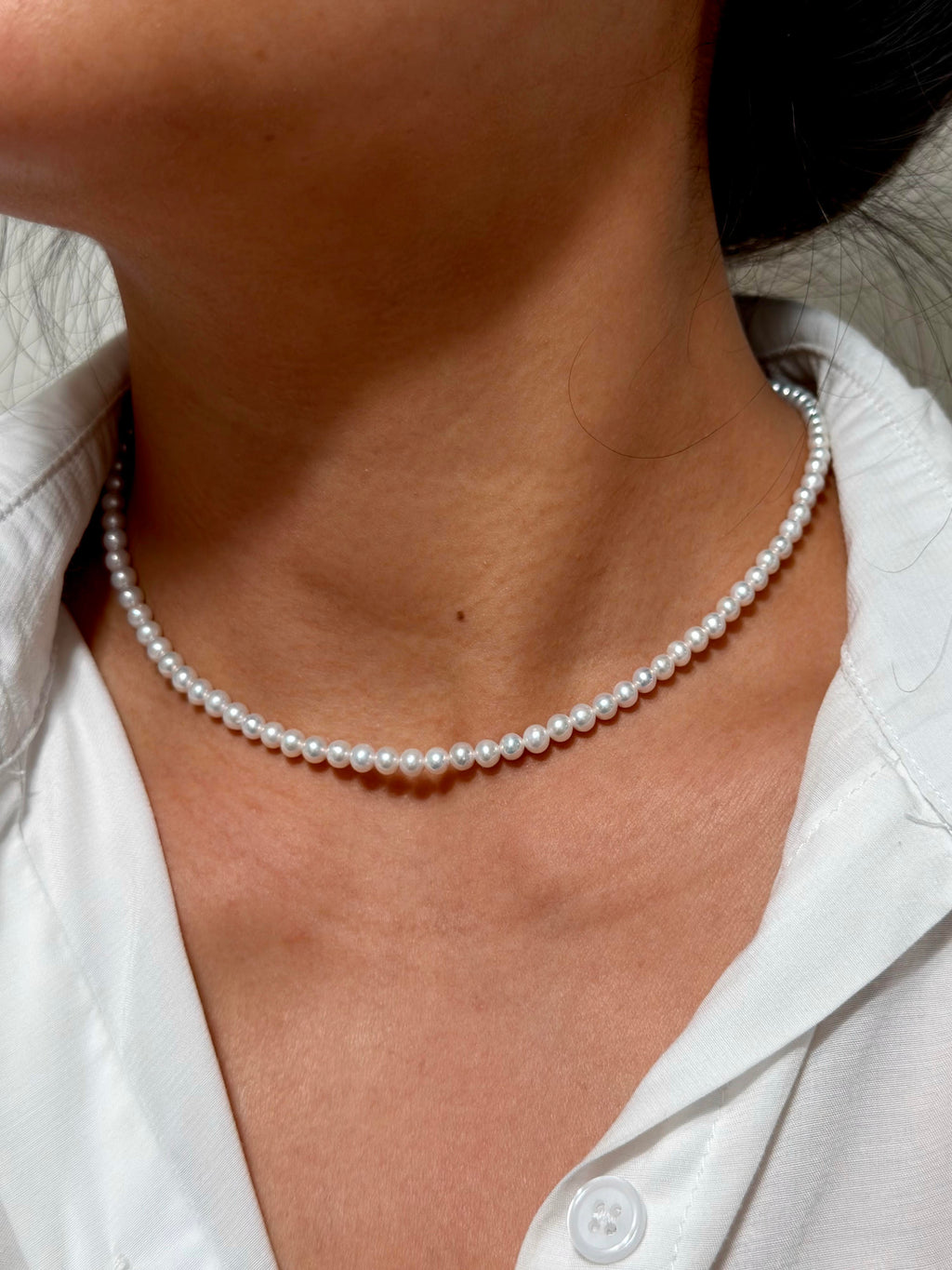 A close-up of a model's neck and chest wearing a delicate single-strand white pearl choker necklace, paired with a white collared shirt.