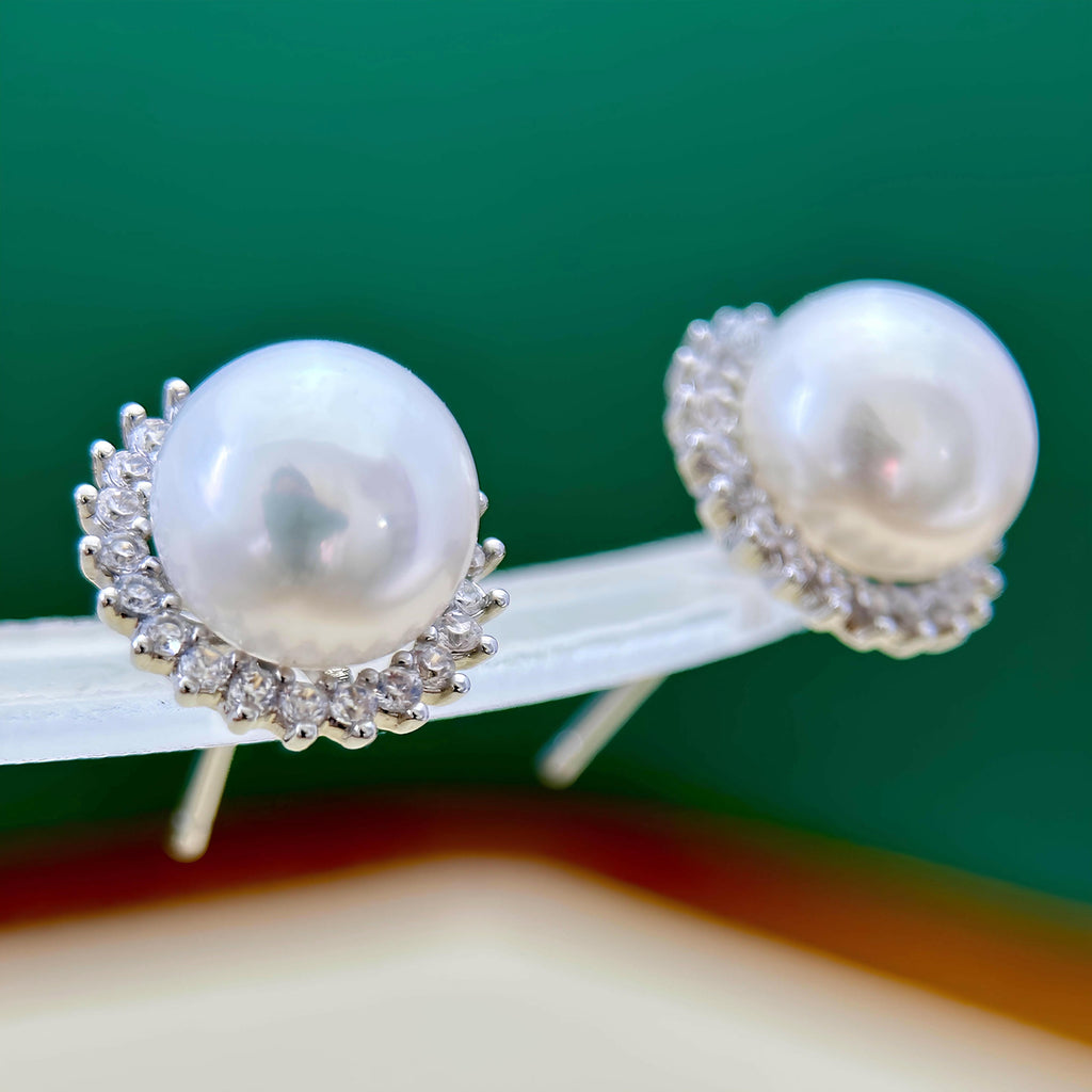 A pair of pearl stud earrings with silver-toned halo settings, displayed on a clear stand against a green background.