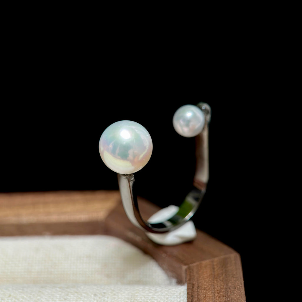 A silver ring featuring two round white pearls, one large and one small, displayed on a wooden stand against a solid black background.