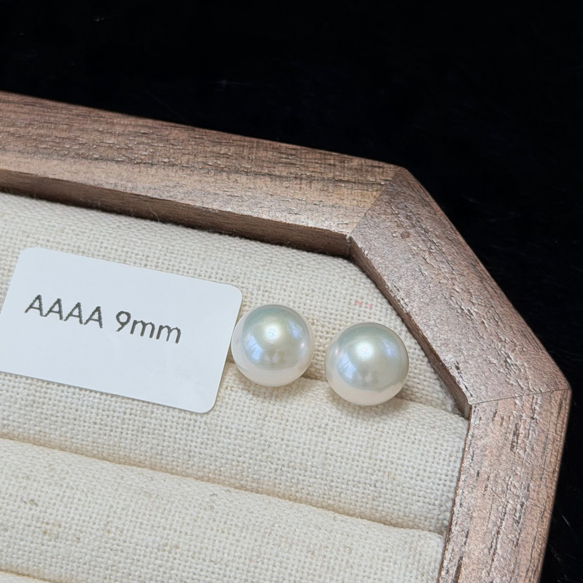 Two round AAAA 9mm freshwater pearl stud earrings displayed on a beige fabric-lined jewelry box with a wooden frame, next to a white size label.