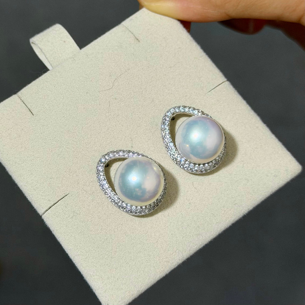 A pair of silver-toned oval stud earrings with large iridescent white pearls and a border of small clear stones, displayed on a cream jewelry card held by a hand.