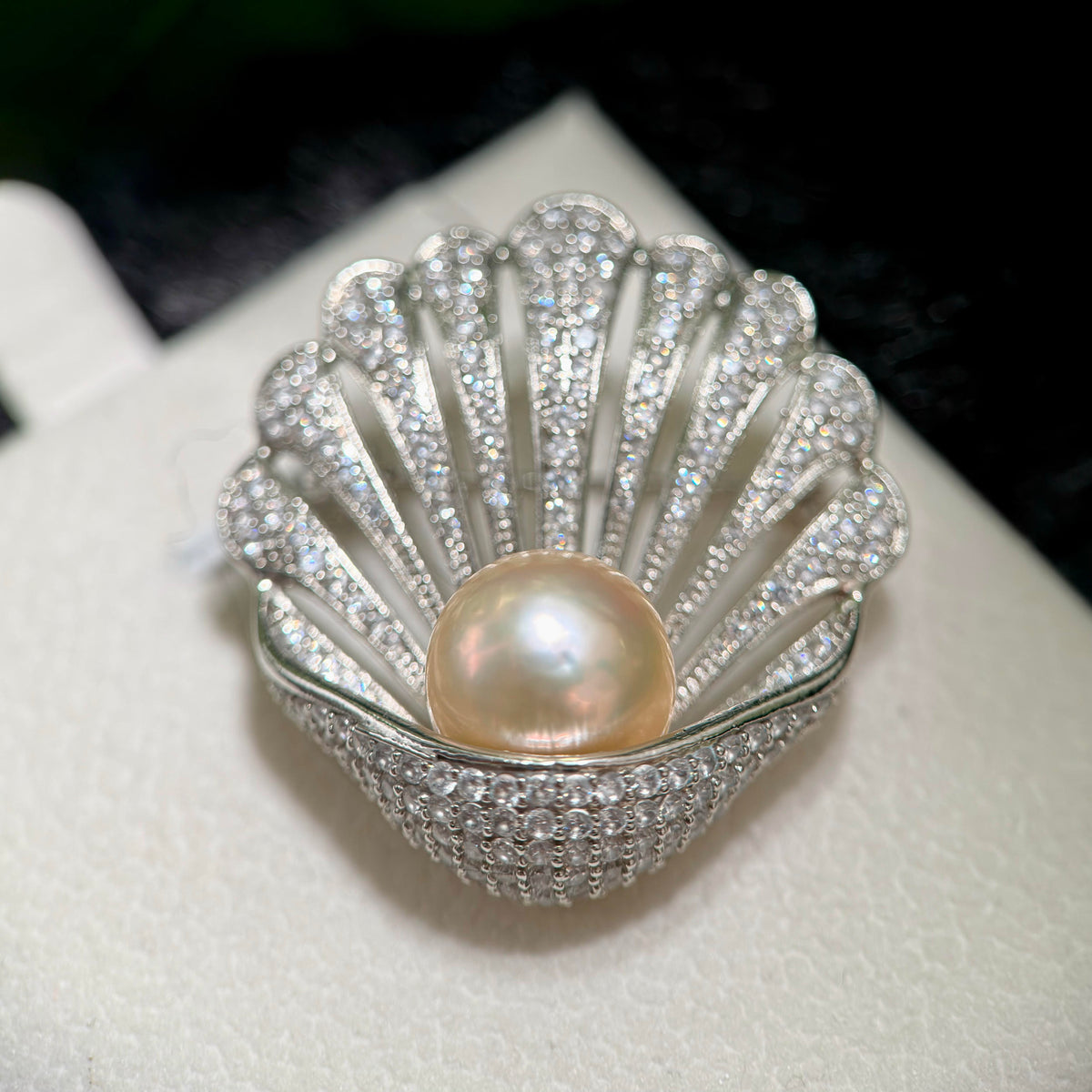 A shell-shaped silver ring brooch or pin featuring a central round pearl and a border of sparkling clear stones, displayed on a white textured surface.