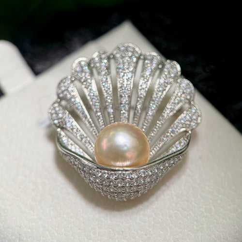 A shell-shaped silver ring brooch or pin featuring a central round pearl and a border of sparkling clear stones, displayed on a white textured surface.