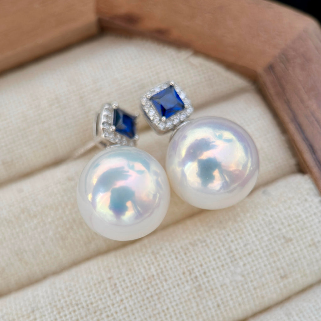 A pair of stud earrings with large iridescent white pearls and square blue gemstones in silver settings, displayed on a cream fabric jewelry cushion inside a wooden box.