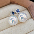 A pair of stud earrings with large iridescent white pearls and square blue gemstones in silver settings, displayed on a cream fabric jewelry cushion inside a wooden box.