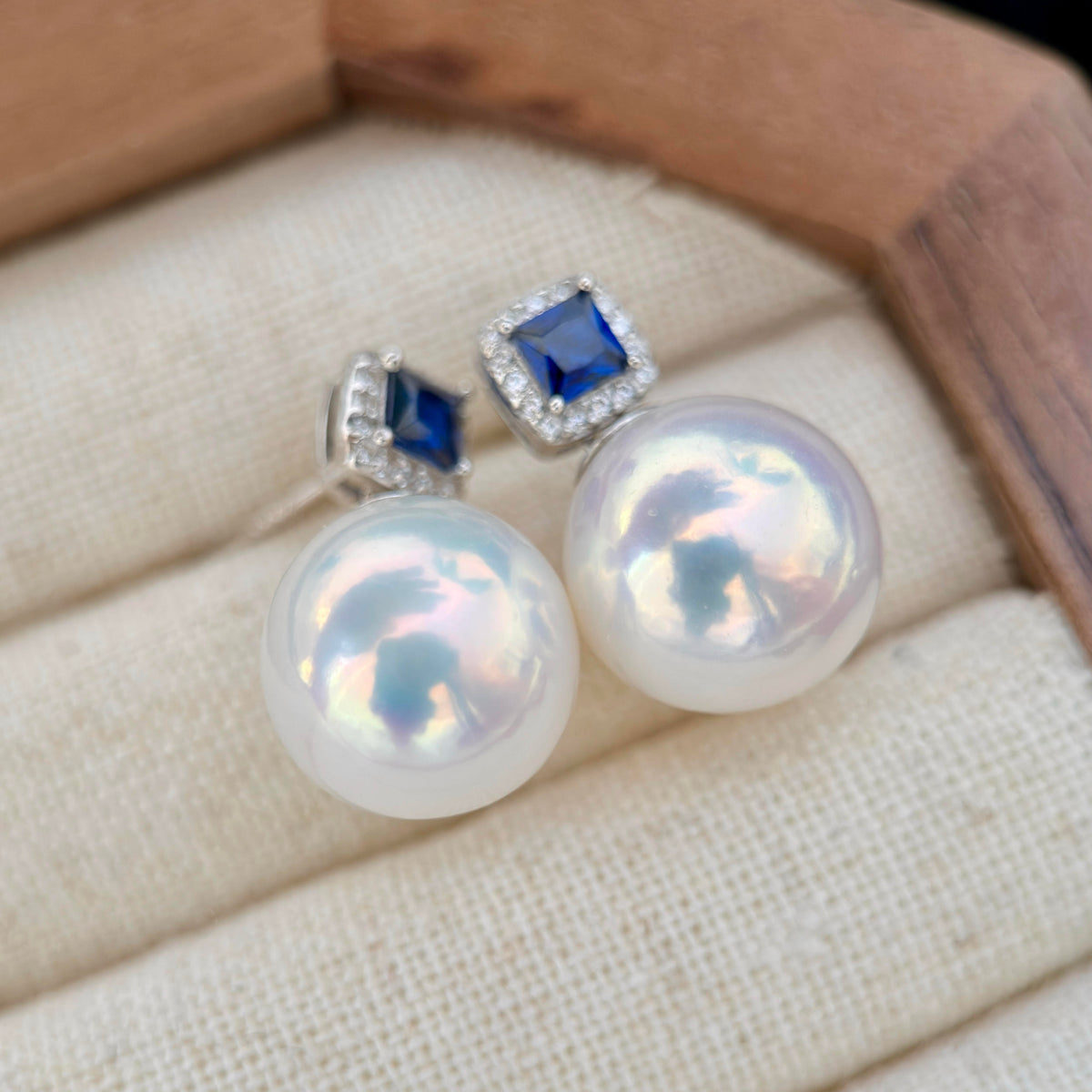 A pair of stud earrings with large iridescent white pearls and square blue gemstones in silver settings, displayed on a cream fabric jewelry cushion inside a wooden box.