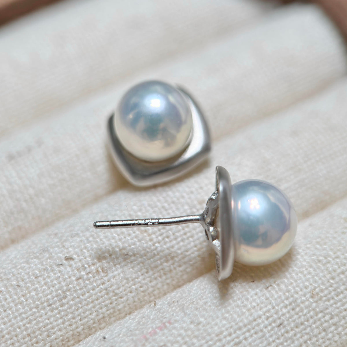 A pair of silver stud earrings with round white freshwater pearls, displayed on a textured beige fabric surface.