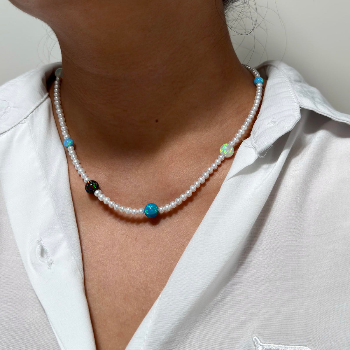 A close-up of a person wearing a beaded necklace with white pearl-like beads, turquoise blue beads, and a small black bead, paired with a white collared shirt.