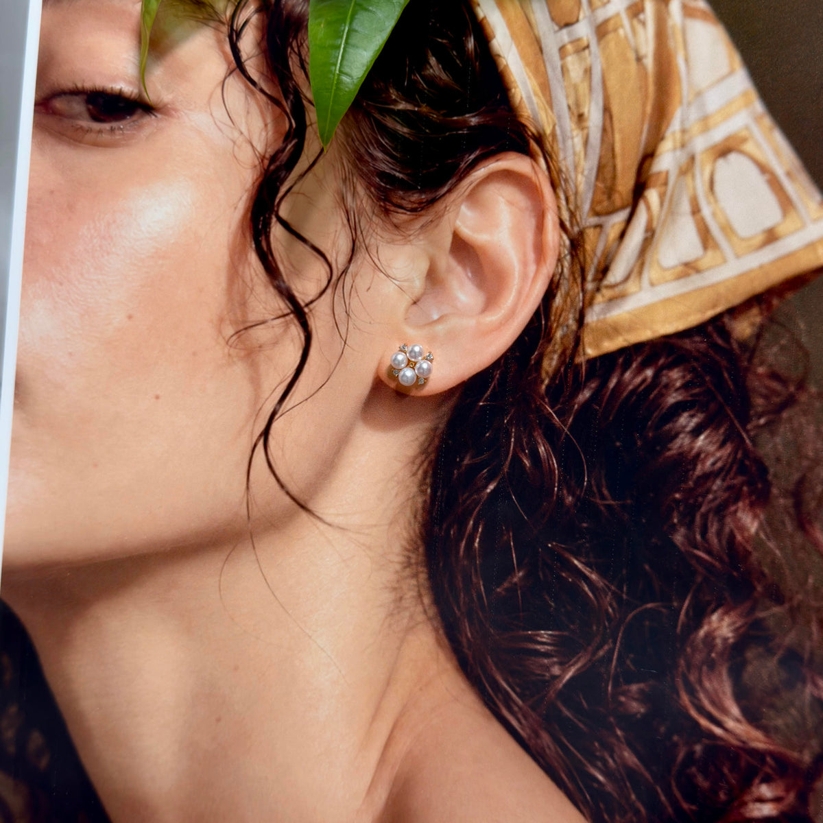 A close-up lifestyle shot of a woman wearing a small, round cluster stud earring with clear gemstones in a silver-toned setting. She has dark curly hair, a patterned beige headscarf, and a green leaf tucked near her eye.